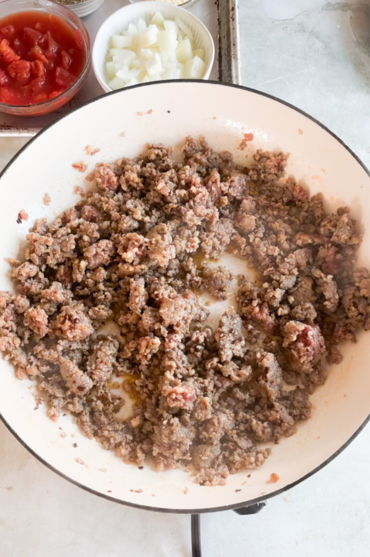 Ground beef is being browned in a white skillet, with bowls of chopped onions and canned tomatoes visible in the background.