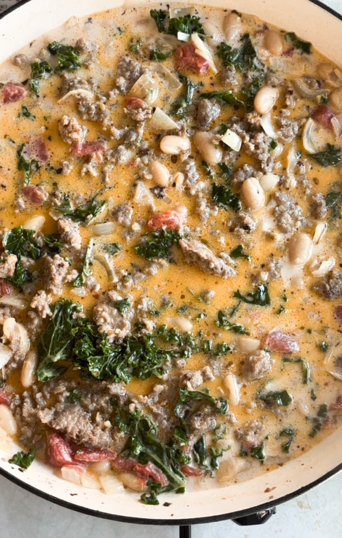 A close-up of a creamy soup in a pot with ground meat, white beans, chopped kale, tomatoes, and onions.