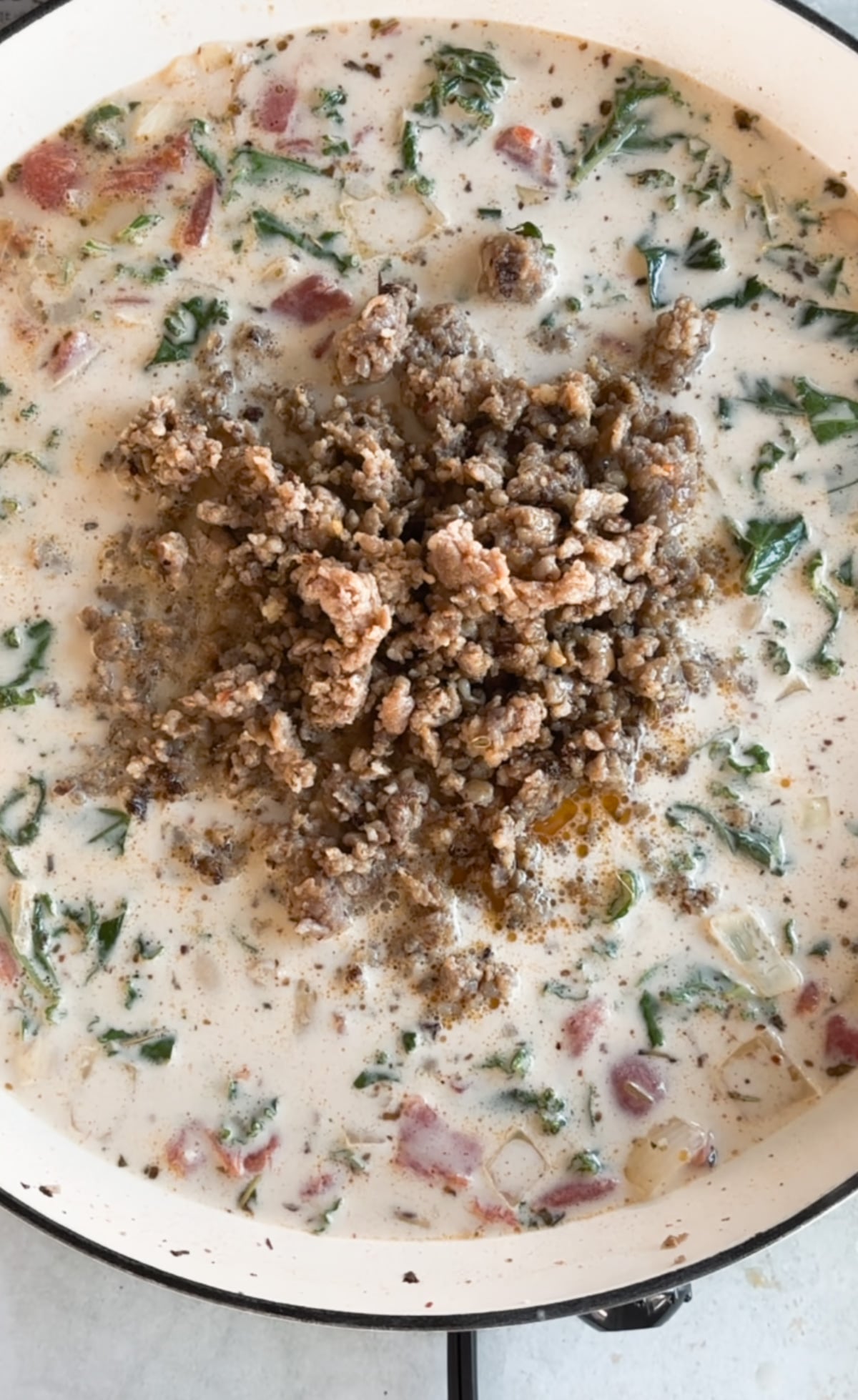 A close-up of a creamy soup in a pot with cooked ground meat, chopped greens, diced tomatoes, and visible spices.