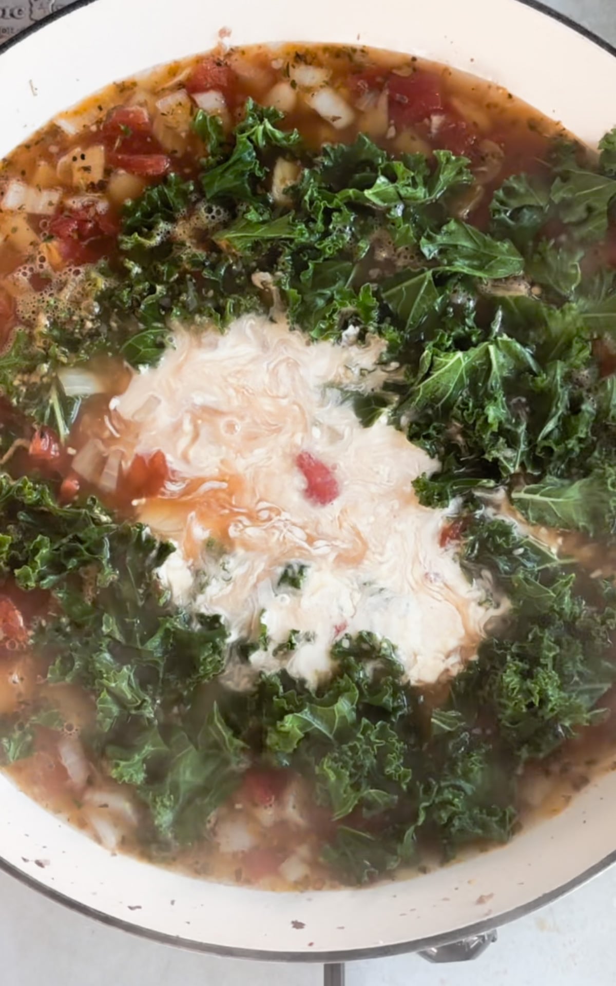 A pot of soup with chopped kale, diced tomatoes, onions, broth, and a swirl of cream simmering on the stove.
