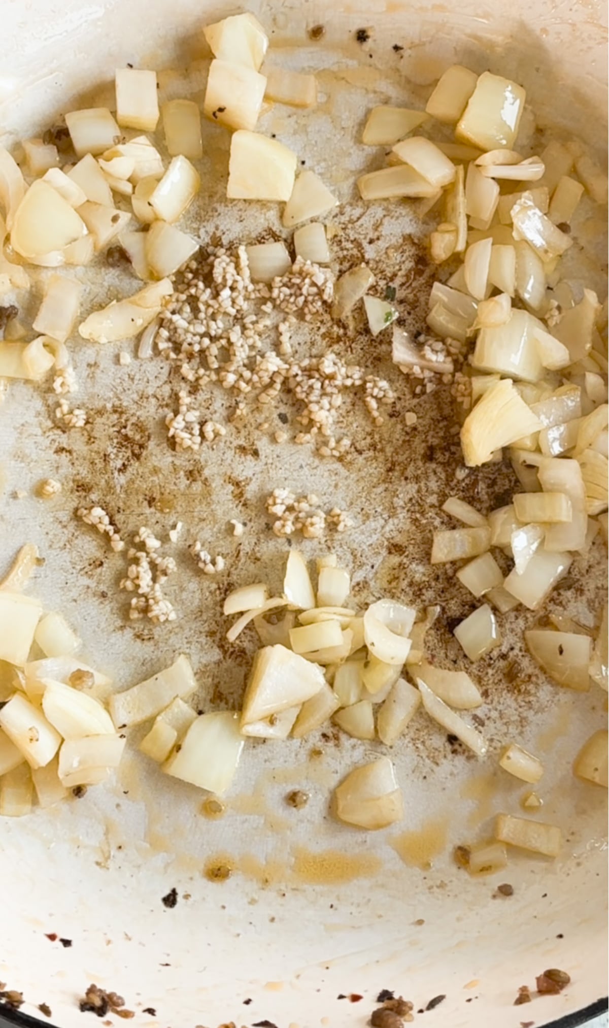 Chopped onions and minced garlic sautéing in a white pan with oil and browned bits.