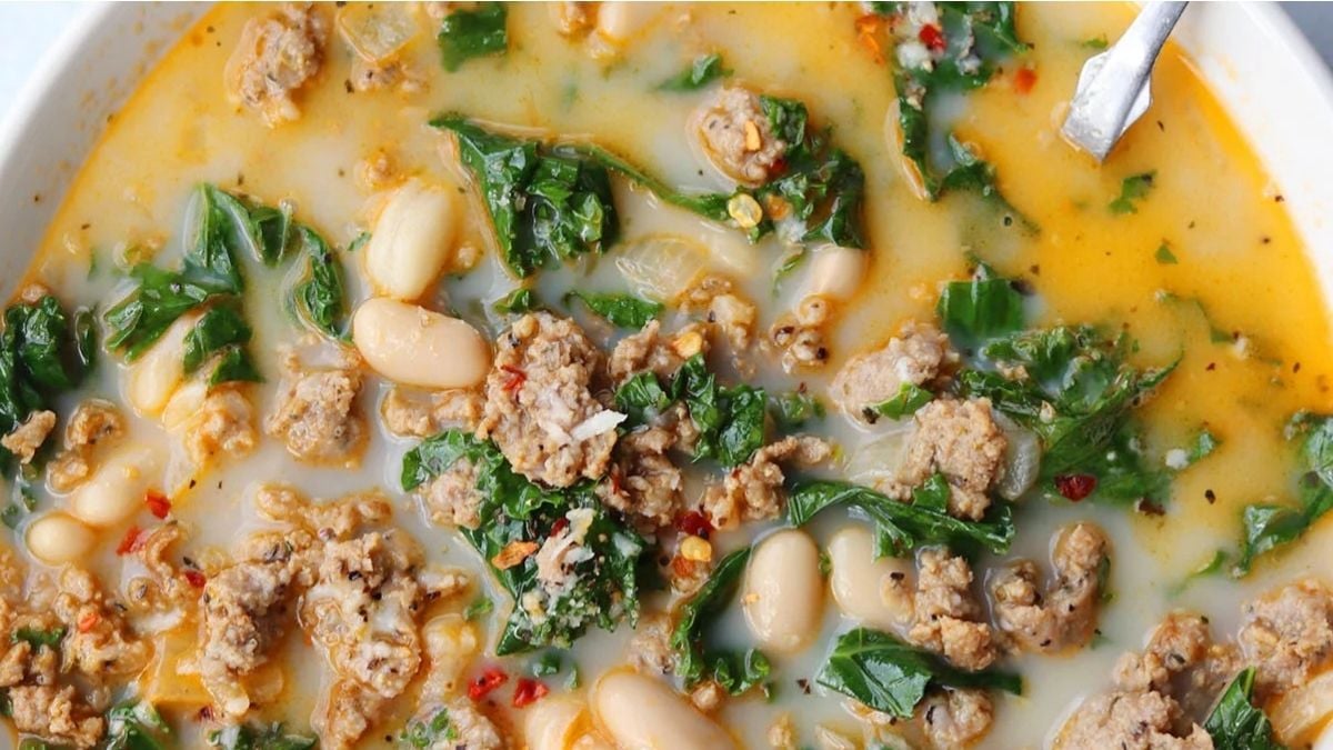 A bowl of creamy soup with ground sausage, white beans, kale, and red pepper flakes, with a spoon in the bowl.