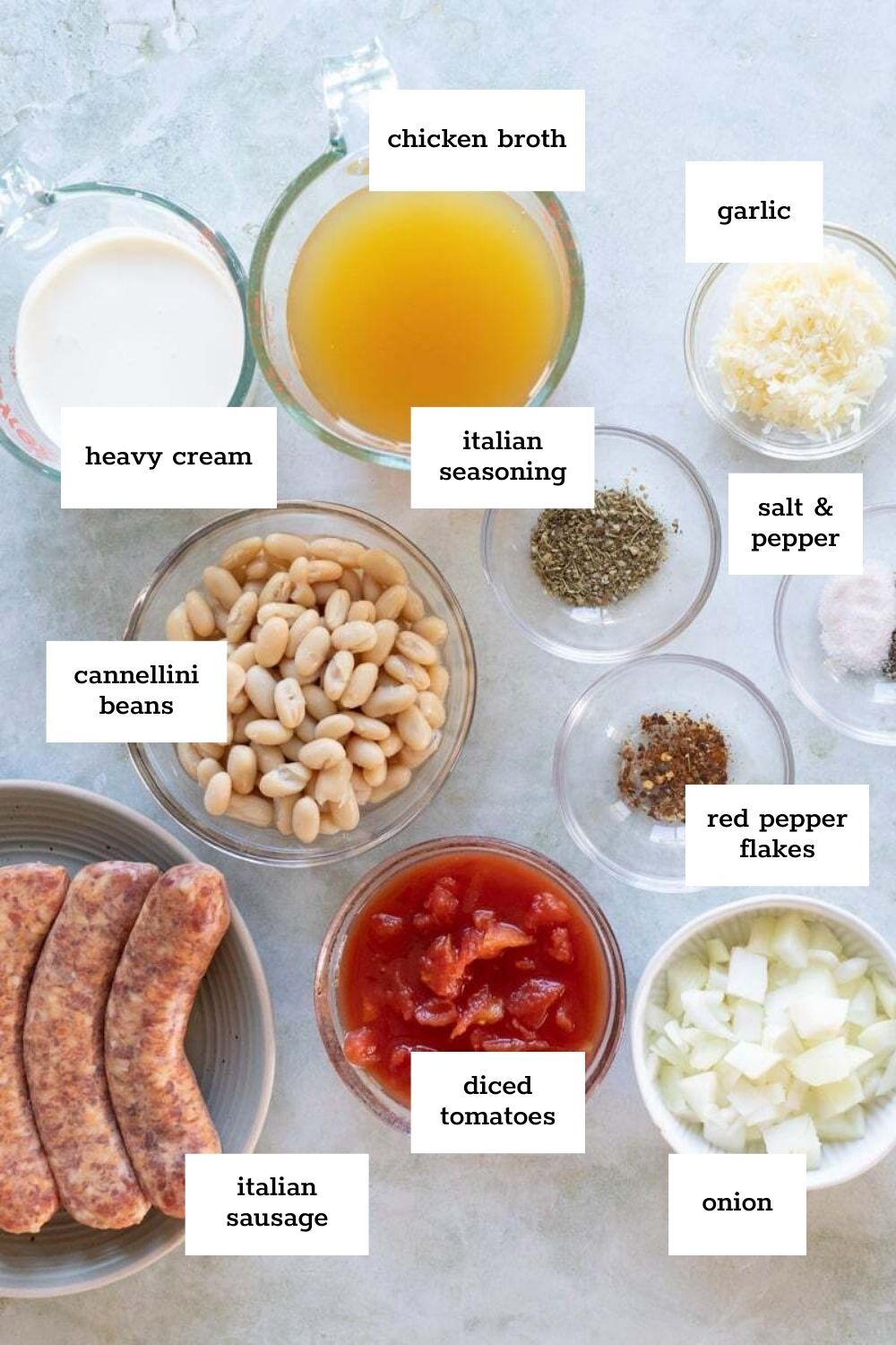 Bowls and cups filled with ingredients for Tuscan bean soup, including heavy cream, chicken broth, garlic, Italian seasoning, cannellini beans, diced tomatoes, onion, and Italian sausage.