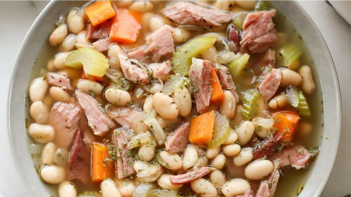 A bowl of ham and bean soup with white beans, ham pieces, sliced carrots, celery, and herbs in broth.
