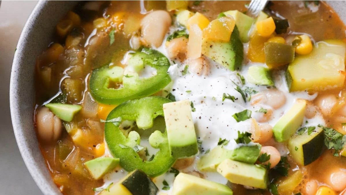 A bowl of vegetable and bean soup topped with sliced jalapeños, diced avocado, chopped herbs, and a dollop of creamy sauce.