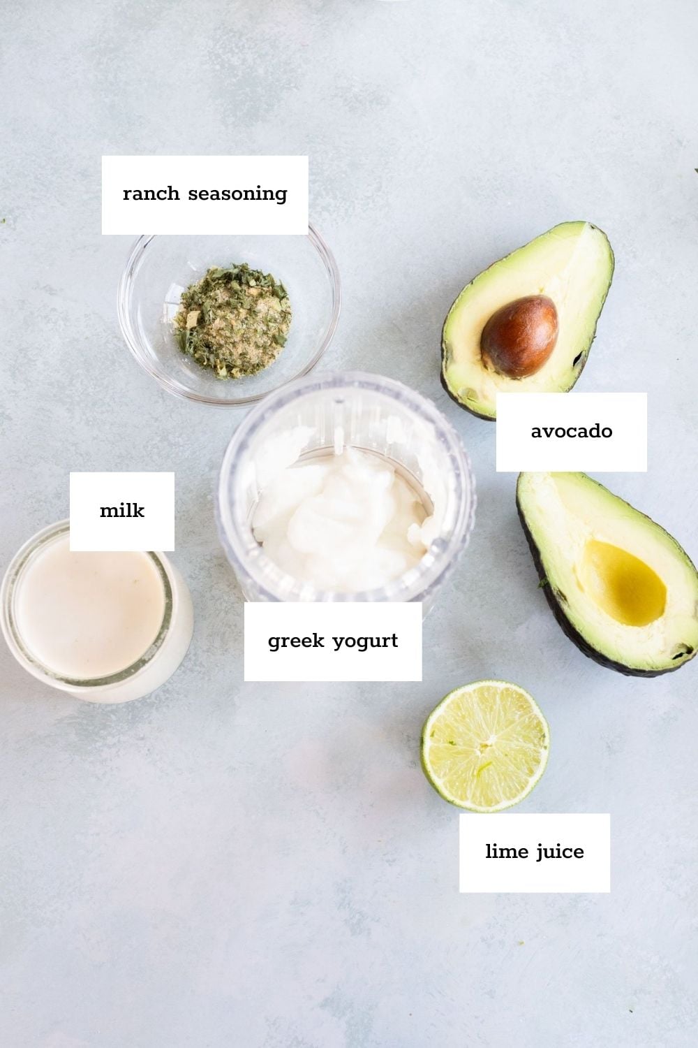 Flat lay of labeled ingredients for a recipe: ranch seasoning, milk, Greek yogurt, avocado halves, and a halved lime with visible juice.