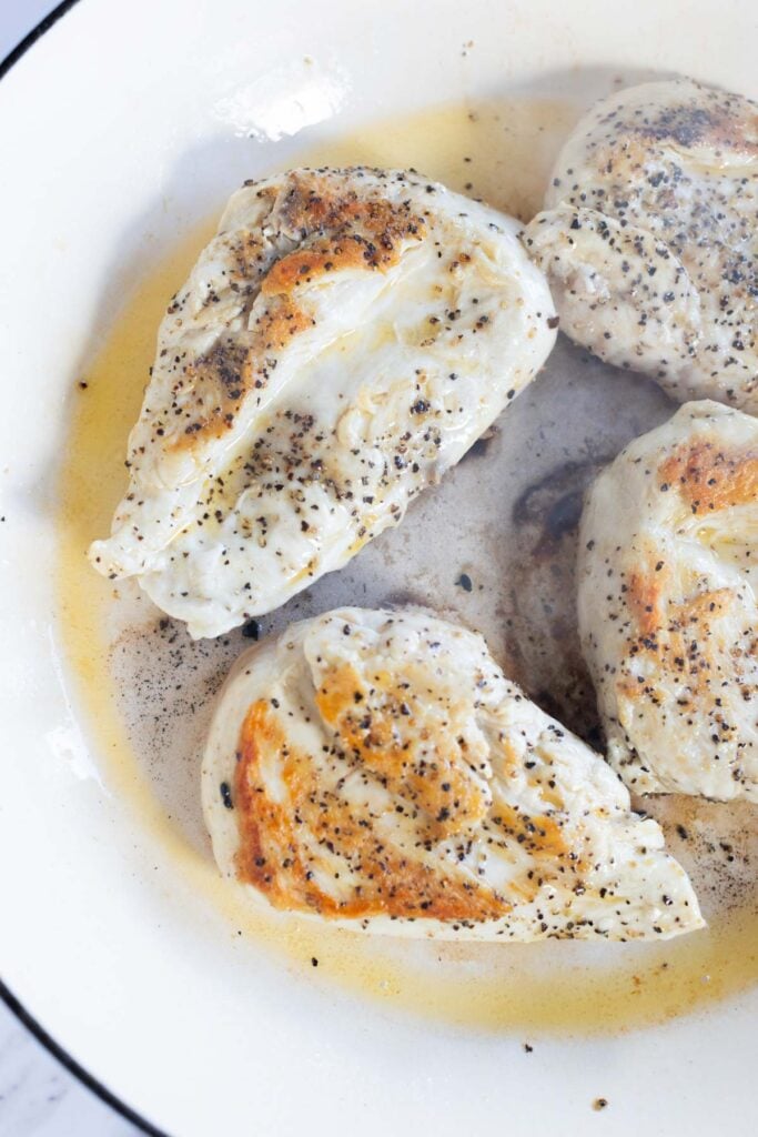 Four cooked chicken breasts seasoned with salt and pepper in a white pan, showing some browning and juices around the meat.