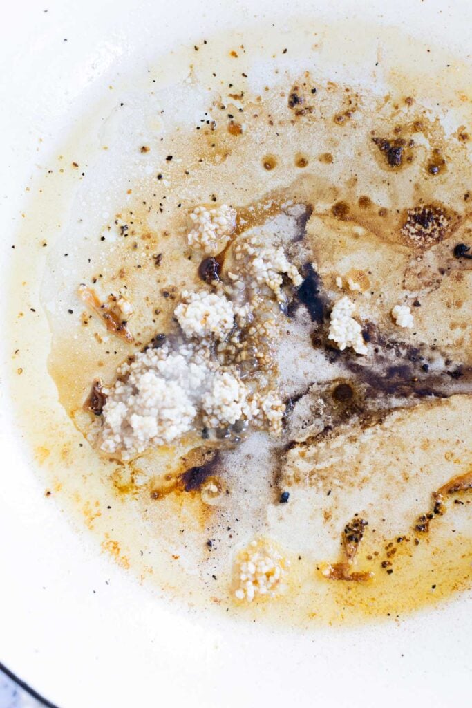 Close-up of a white pan with browned bits, oil, and clumps of undissolved flour and minced garlic.