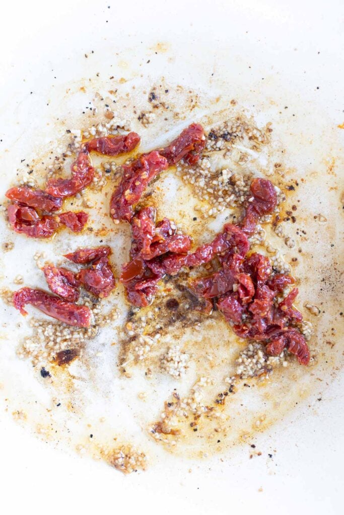 Chopped sun-dried tomatoes and minced garlic sautéing in oil and spices in a white pan.