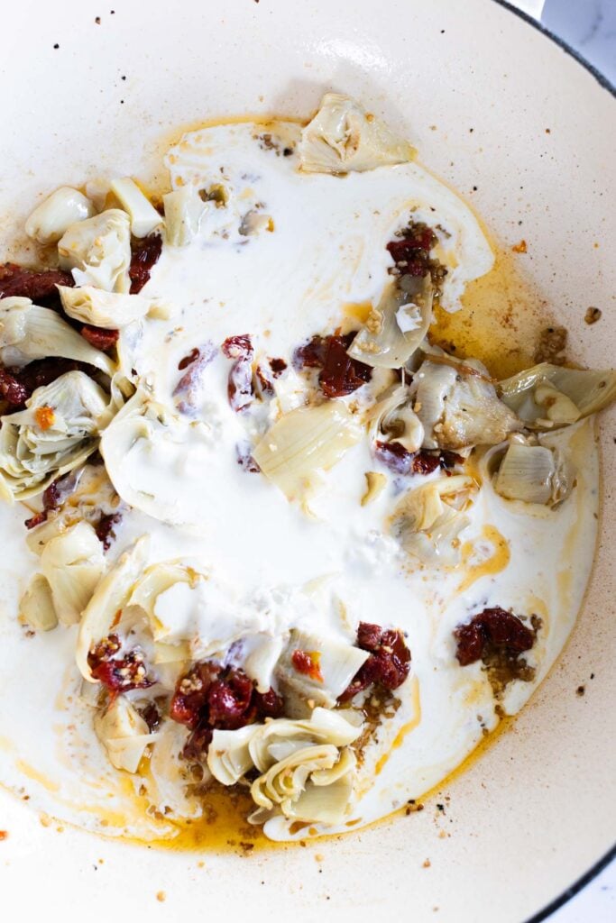 Artichoke hearts, sun-dried tomatoes, and cream are being cooked together in a white pan.
