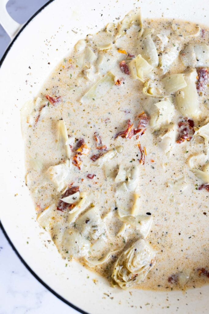 Creamy sauce with artichoke hearts and sun-dried tomatoes simmering in a white pan.