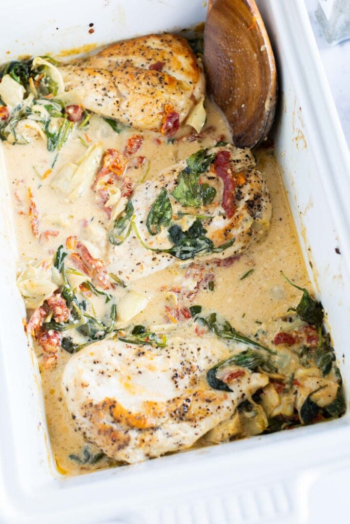 Baked Tuscan chicken in a creamy sauce with spinach, sun-dried tomatoes, and artichokes in a white ceramic baking dish, with a wooden spoon in the corner.