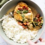 A bowl with white rice and creamy, baked Tuscan chicken breast topped with spinach and sun-dried tomatoes, with a fork and a hand holding the bowl.