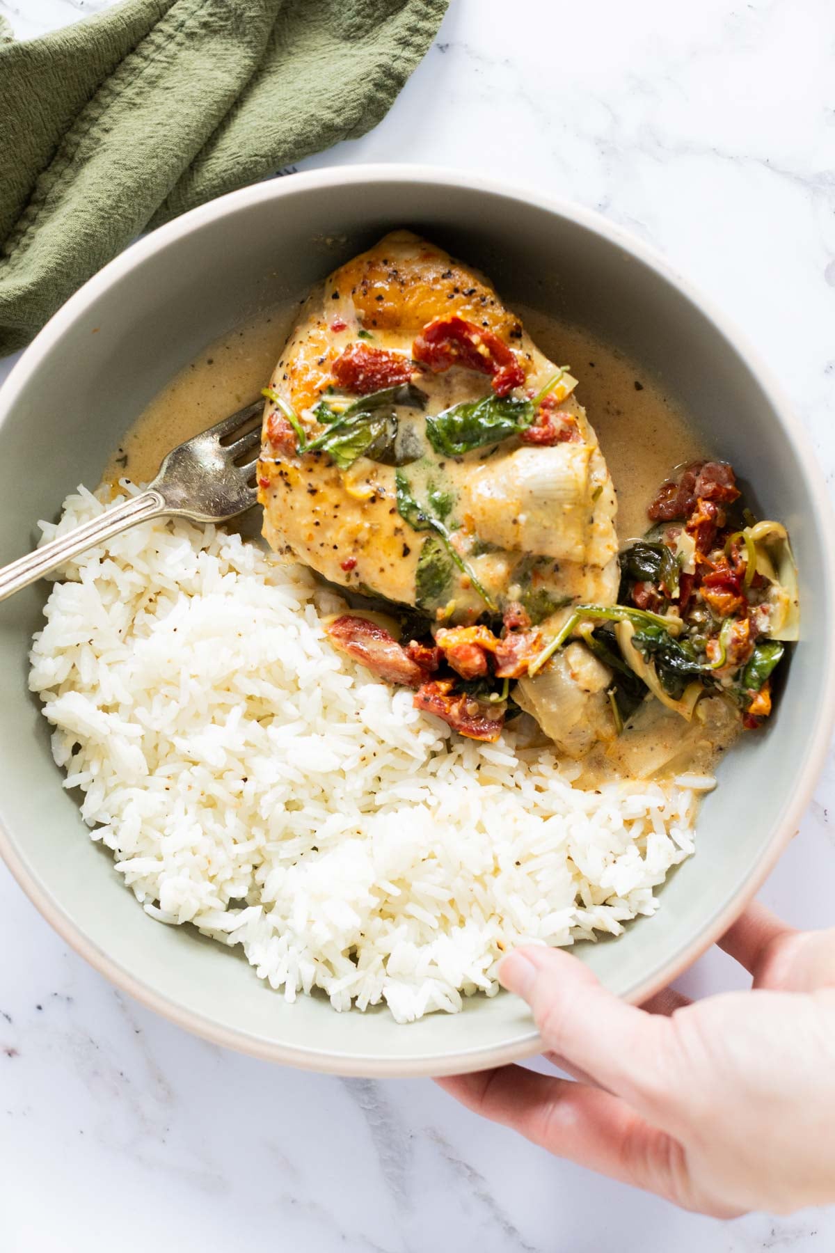 A bowl with white rice and creamy, baked Tuscan chicken breast topped with spinach and sun-dried tomatoes, with a fork and a hand holding the bowl.