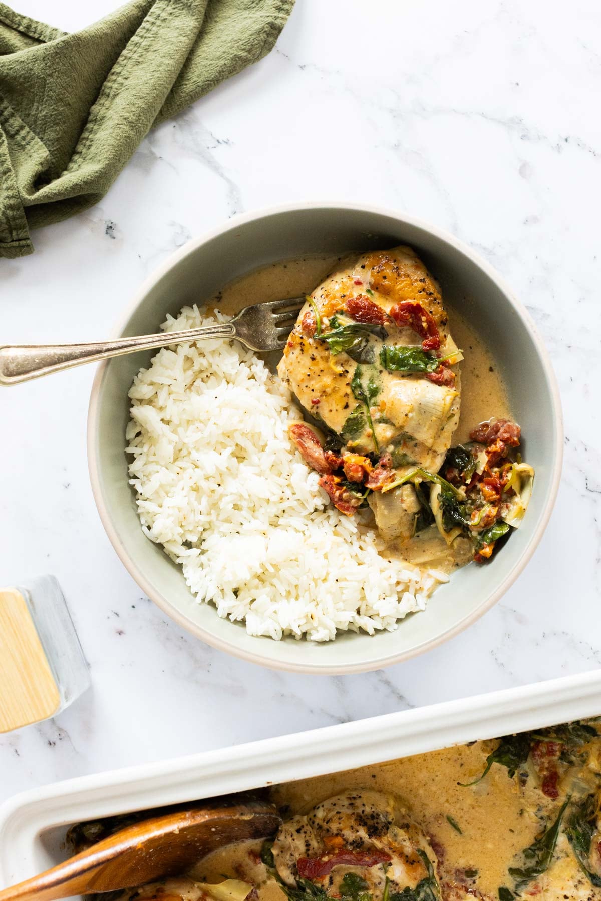 A bowl containing white rice and a piece of creamy, saucy Baked Tuscan chicken with spinach and tomatoes, with a fork placed on the side.