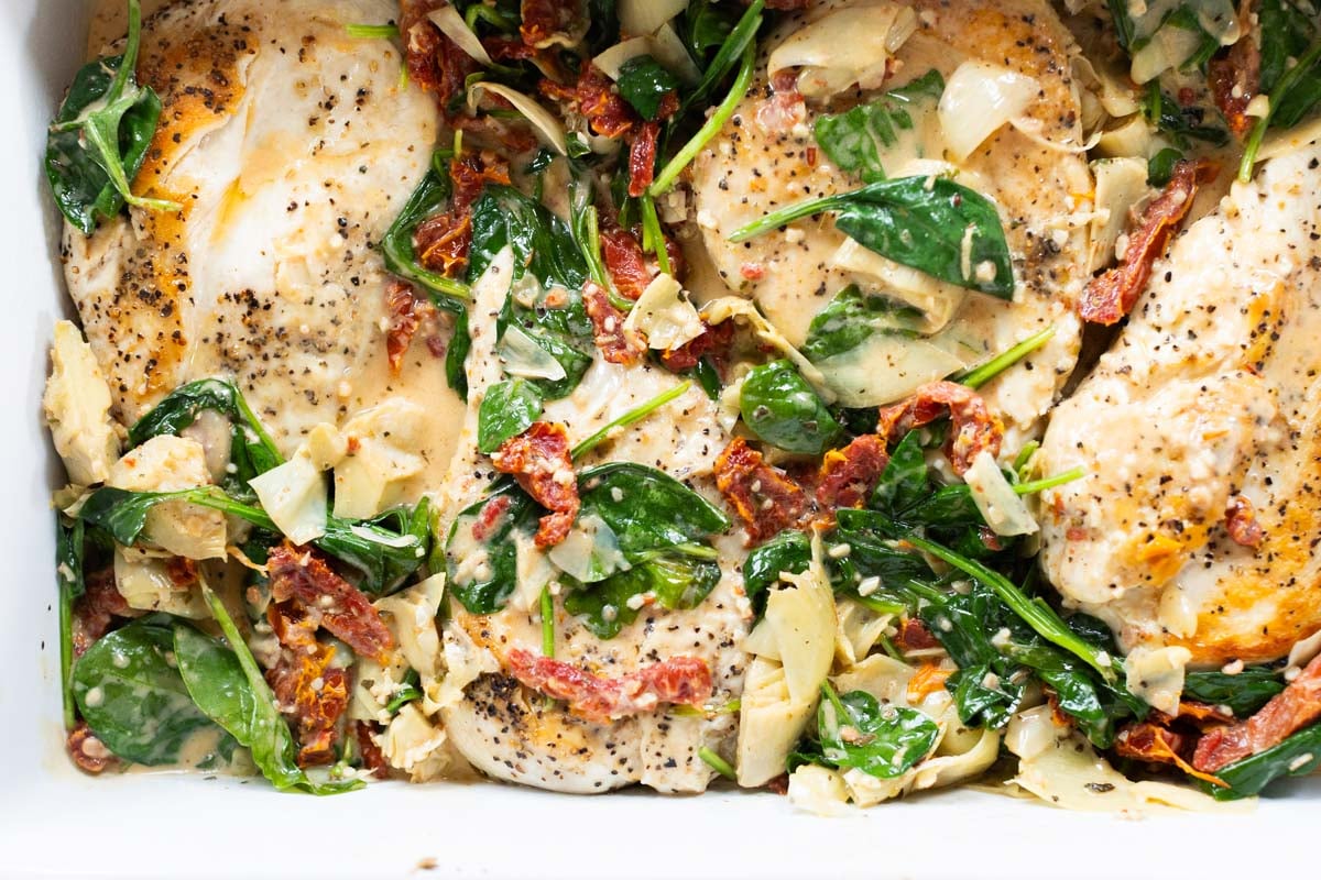 Baked Tuscan chicken breasts with spinach, sun-dried tomatoes, artichoke hearts, and a creamy sauce in a white baking dish.