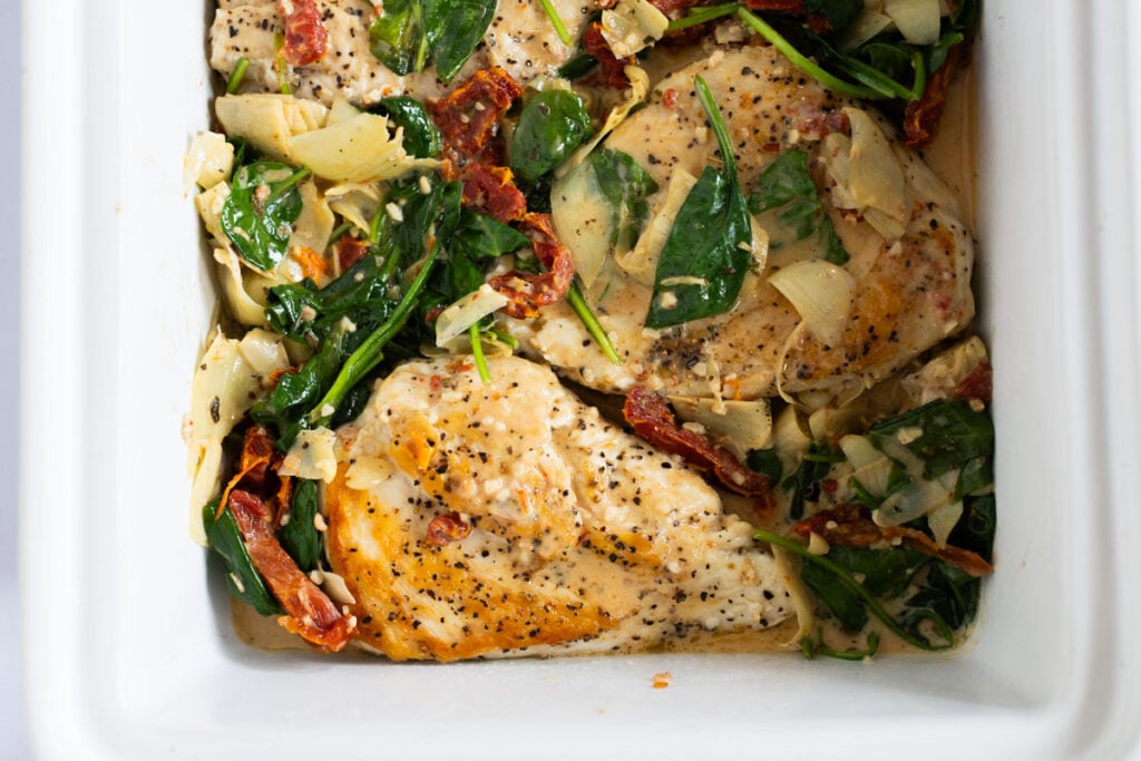 Cooked chicken breasts with spinach, sun-dried tomatoes, and artichokes in a creamy sauce, served in a white baking dish.