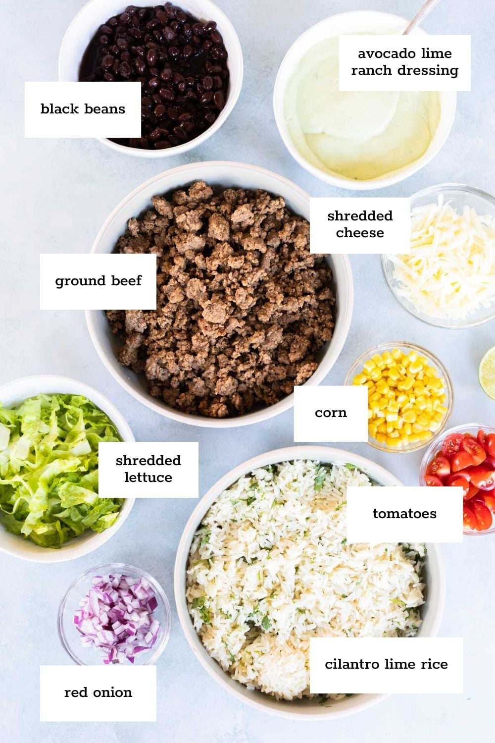 Labeled bowls of black beans, cooked ground beef, shredded cheese, corn, tomatoes, shredded lettuce, red onion, cilantro lime rice, and avocado lime ranch dressing arranged on a table.