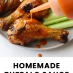 Buffalo sauce being poured onto baked chicken drumsticks, with celery sticks on the plate in the background.