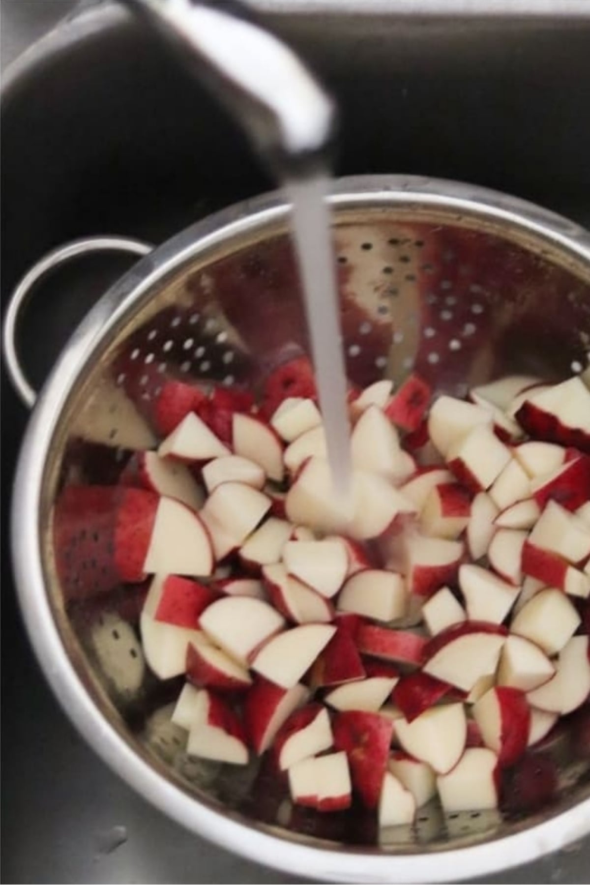 Chopped potatoes in a metal colander being rinsed with running tap water under a kitchen faucet.