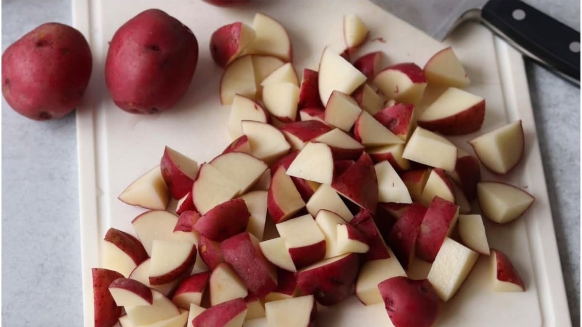 Chopped red potatoes on a white cutting board, with a knife and whole red potatoes in the background.