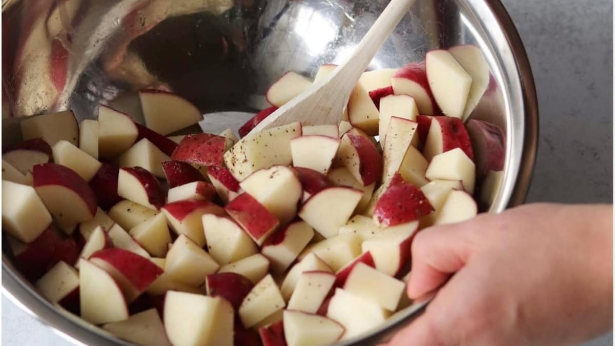A hand stirs chopped red potatoes with a wooden spoon in a large metal mixing bowl.