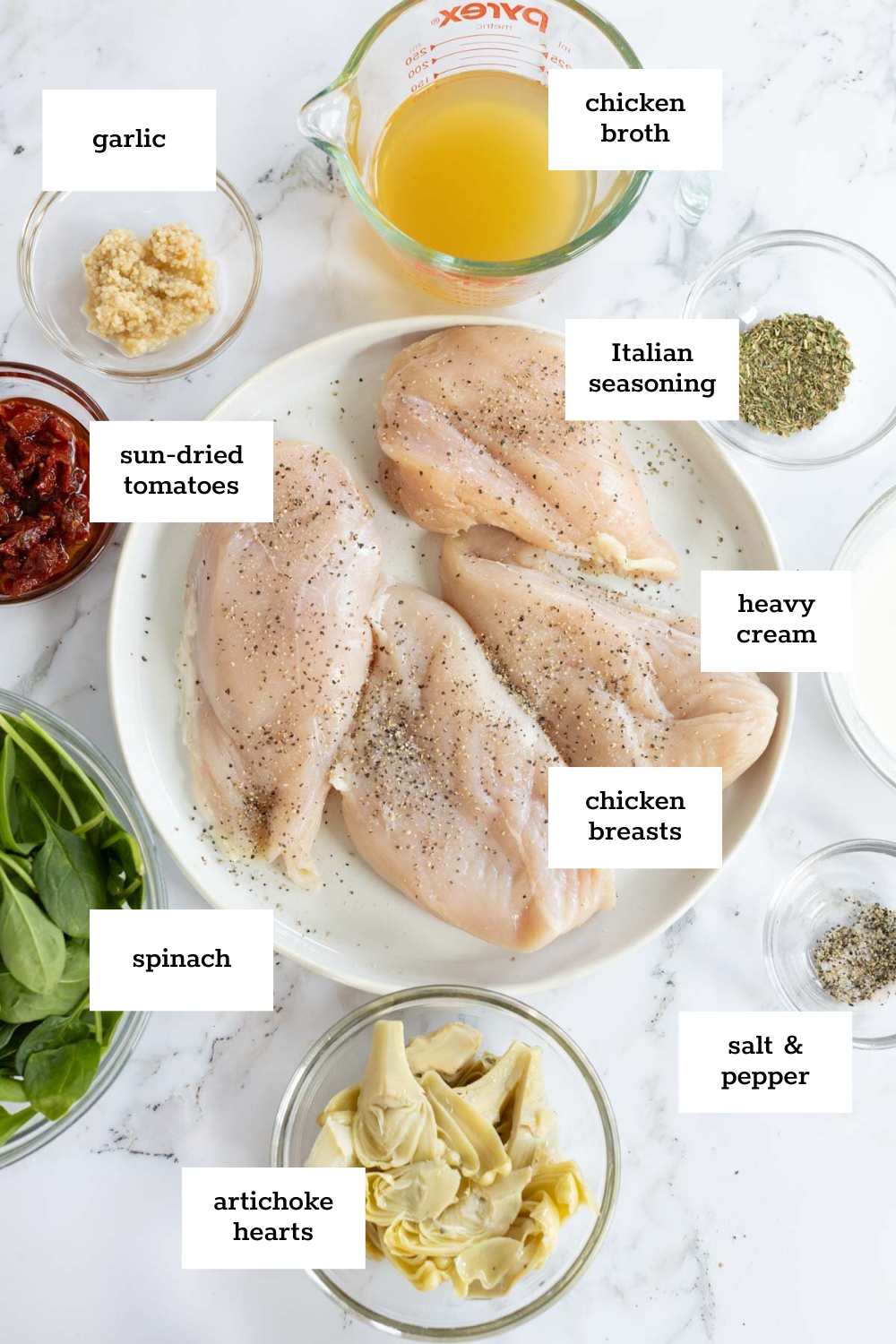 Labeled ingredients for a recipe arranged on a counter, including chicken breasts, Italian spices, sun-dried tomatoes, artichoke hearts, spinach, heavy cream, garlic, and chicken broth.