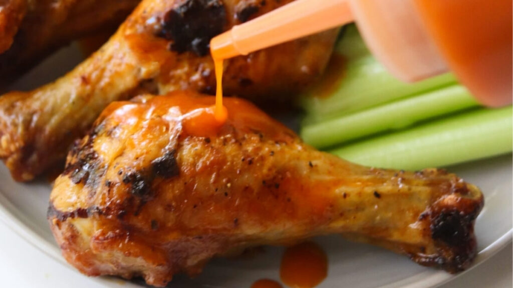 A grilled chicken drumstick is being topped with orange buffalo wing sauce recipe next to several celery sticks on a white plate.
