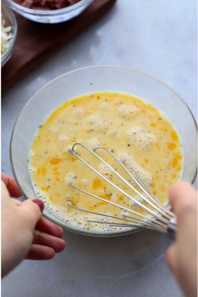 A person whisking eggs and milk in a glass bowl on a light surface, preparing a frittata mixture for cooking.