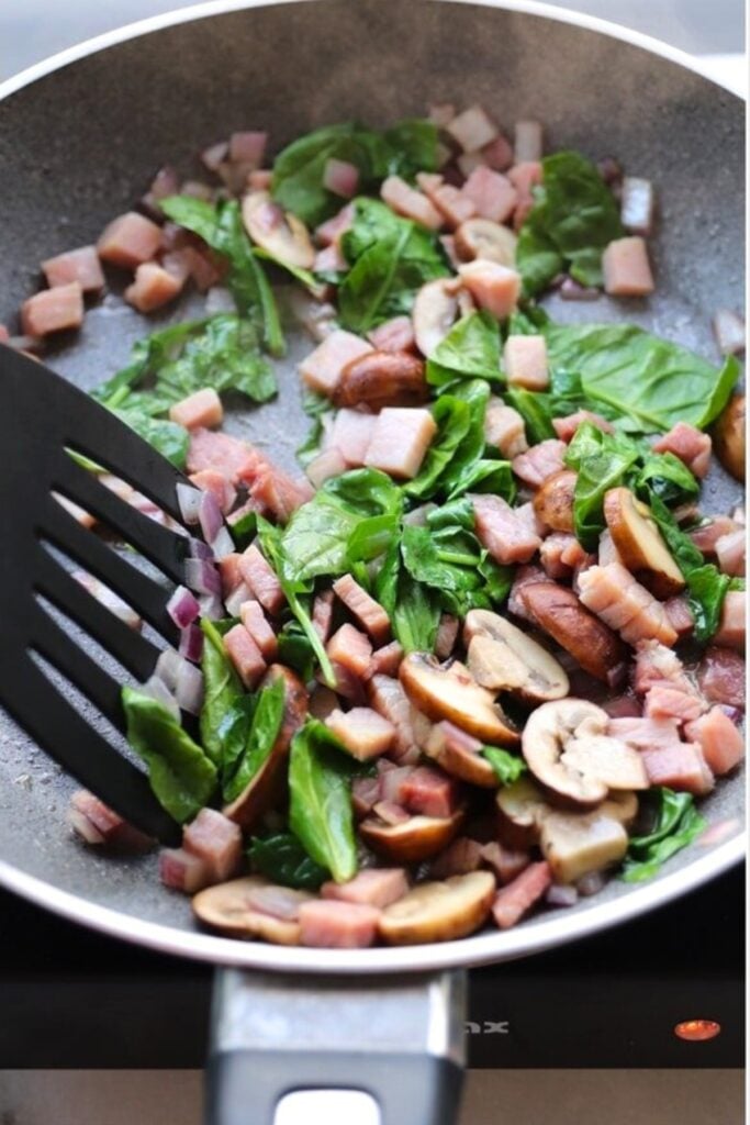A frying pan contains chopped ham, spinach leaves, sliced mushrooms, and red onions being sautéed with a black spatula.