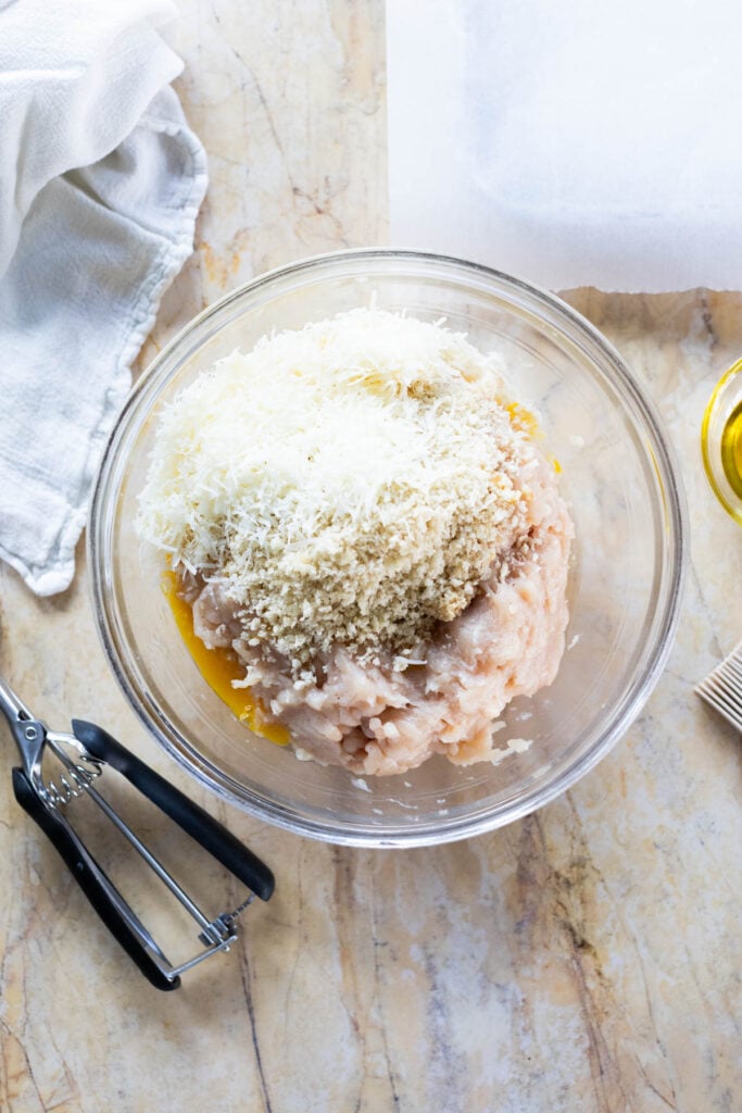 A glass bowl with ground chicken, breadcrumbs, grated cheese, and egg mixture on a marble countertop, next to a cookie scoop and a folded white cloth.