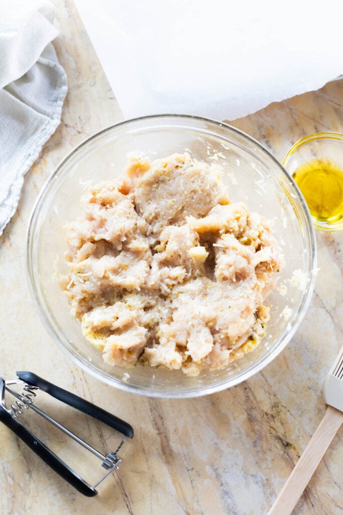 A glass bowl of raw meatball mixture on a marble surface, with a small dish of olive oil, a cookie scoop, and a spatula nearby.