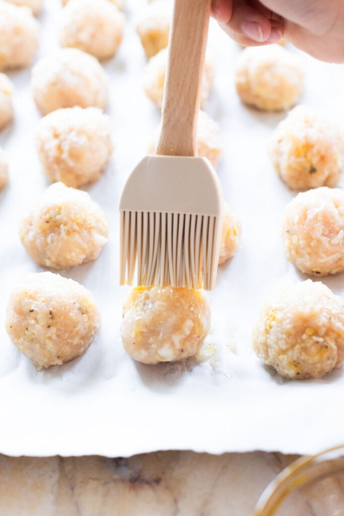 A hand uses a pastry brush to coat uncooked chicken parmesan meatballs with egg wash on a parchment-lined baking sheet.