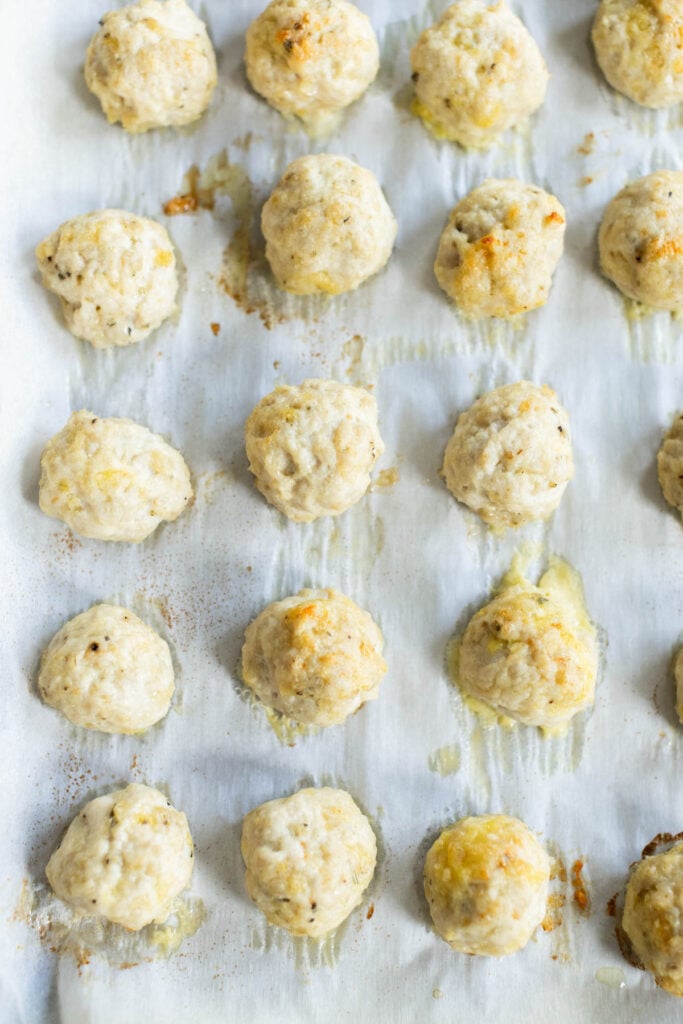 Rows of baked chicken parmesan meatballs on a parchment-lined baking sheet, with some spots of melted cheese and browned edges visible.