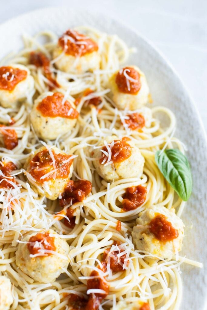 A plate of spaghetti topped with chicken parmesan meatballs, tomato sauce, grated cheese, and a fresh basil leaf on the side.