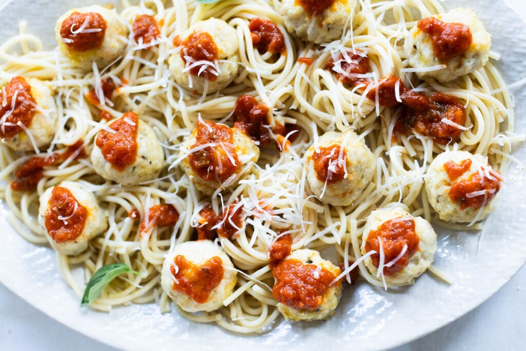 A plate of spaghetti topped with chicken parmesan meatballs, tomato sauce, and grated cheese, garnished with fresh basil leaves.