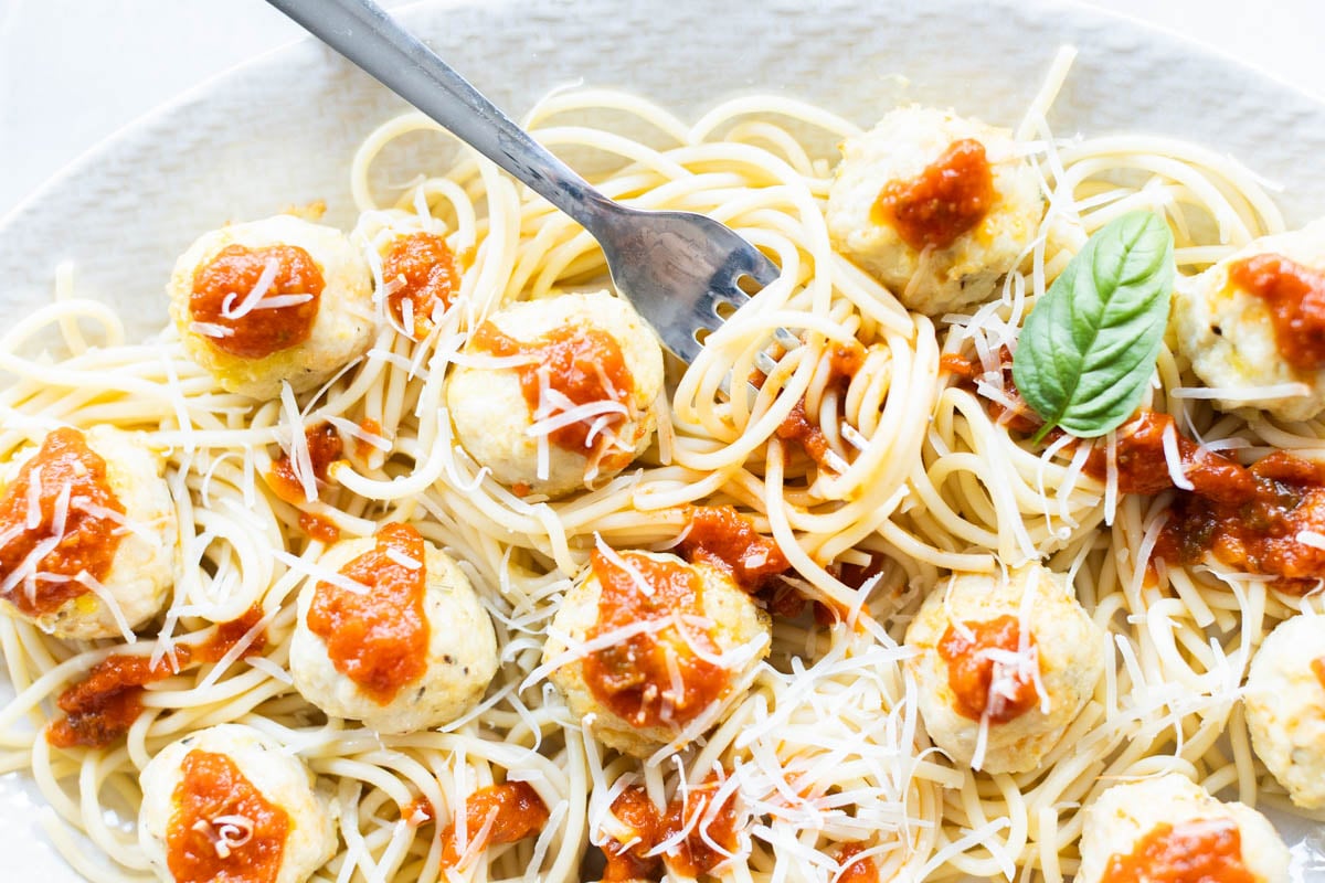 Plate of spaghetti topped with chicken parmesan meatballs, marinara sauce, grated cheese, and a basil leaf, with a fork resting on the noodles.