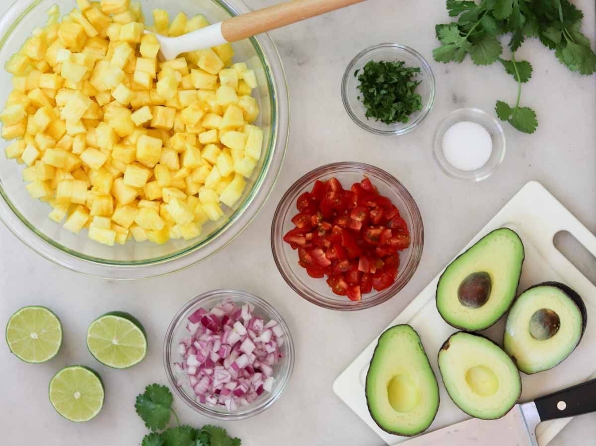 Chopped pineapple, avocado halves, diced tomatoes, red onion, cilantro, lime halves, salt, and a knife arranged on a countertop for food preparation.