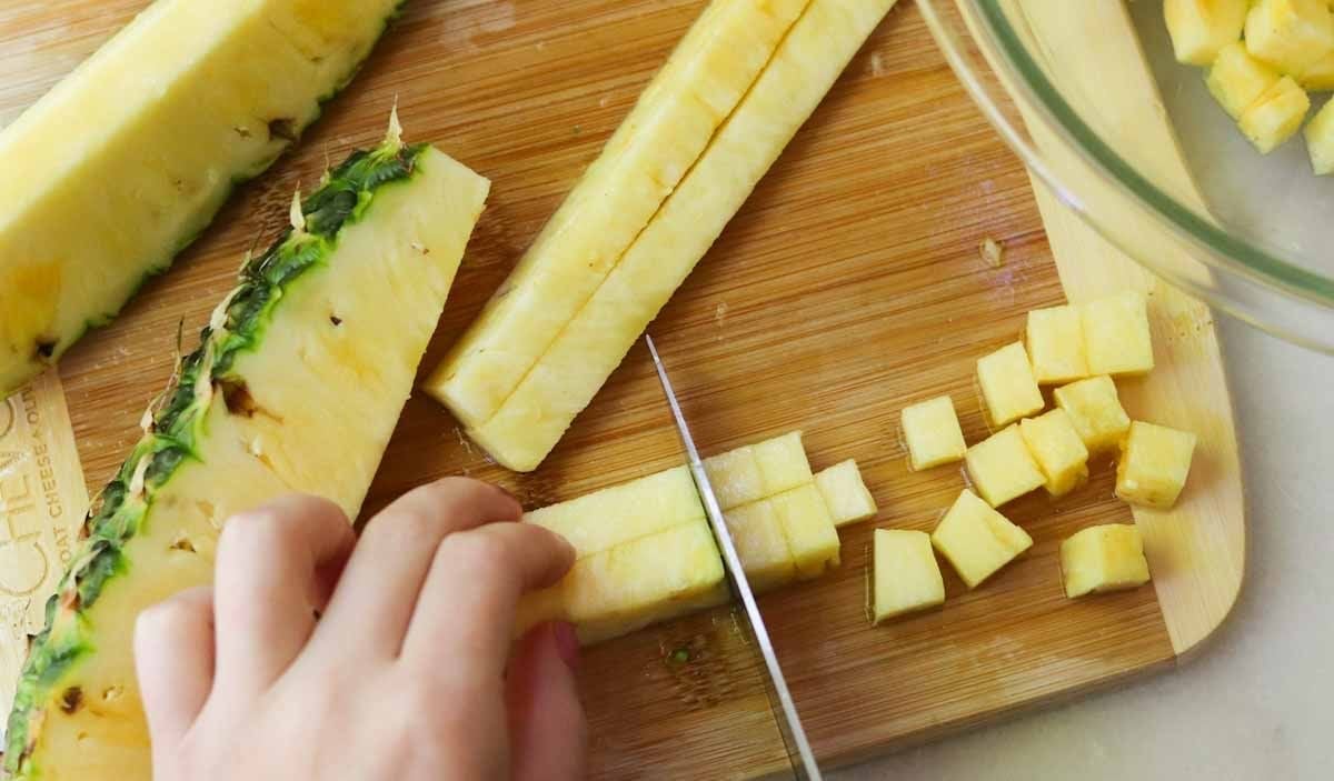 A hand slices fresh pineapple into small cubes on a wooden cutting board, with pineapple pieces and a glass bowl nearby.