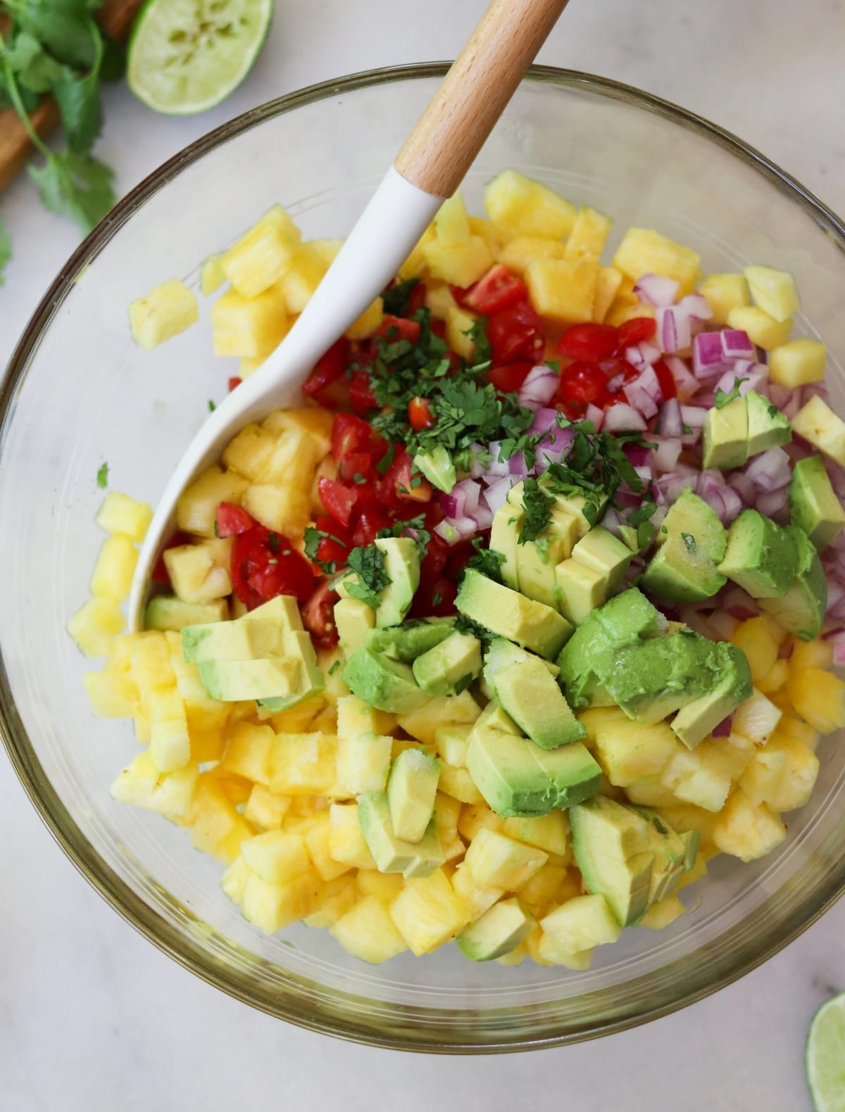 A glass bowl filled with chopped pineapple, avocado, tomato, red onion, and cilantro, with a white and wooden spoon resting inside.