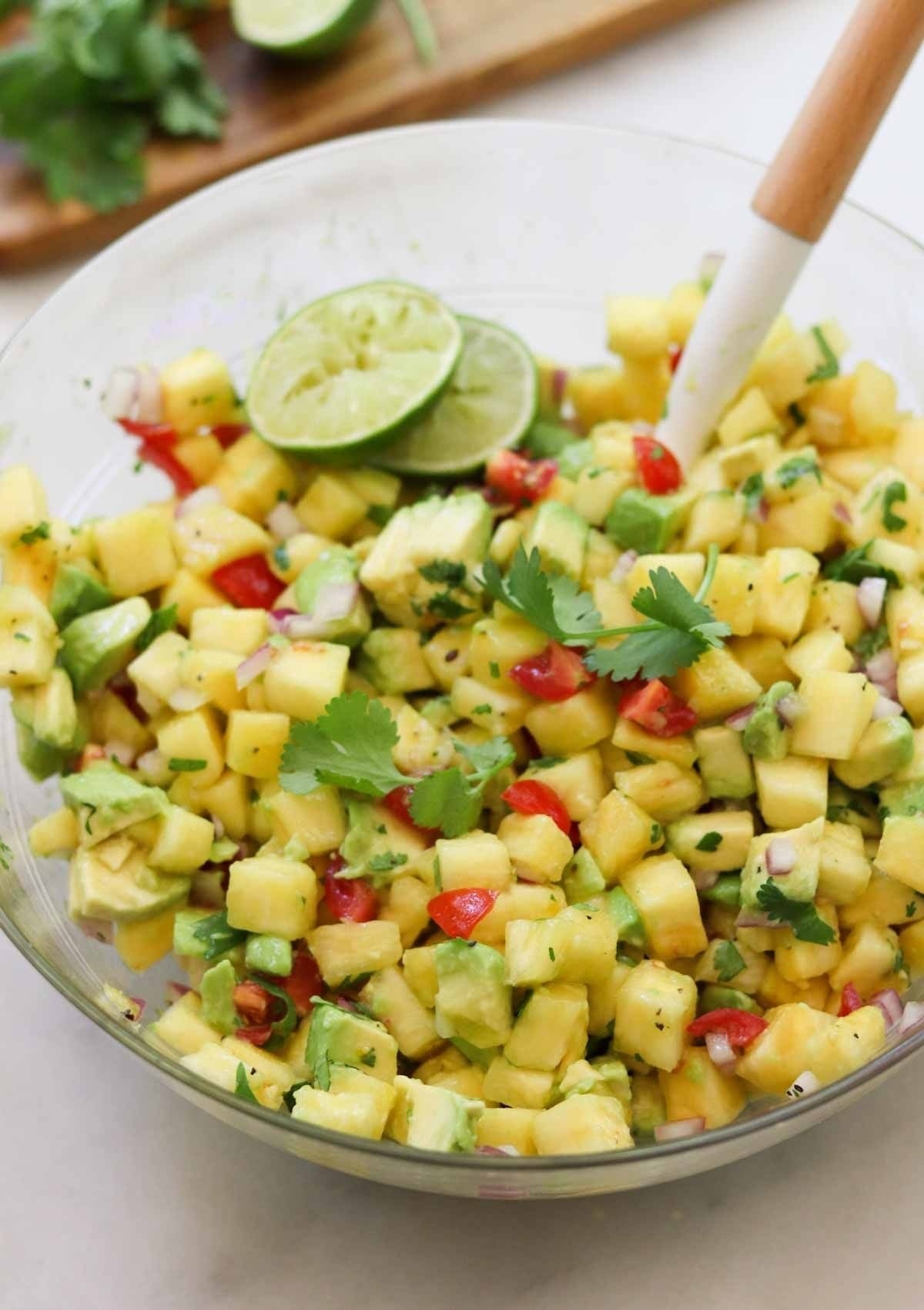 A glass bowl filled with pineapple pico de gallo, containing diced pineapple, avocado, red onion, tomato, cilantro, and lime slices, with a white spoon in the bowl.