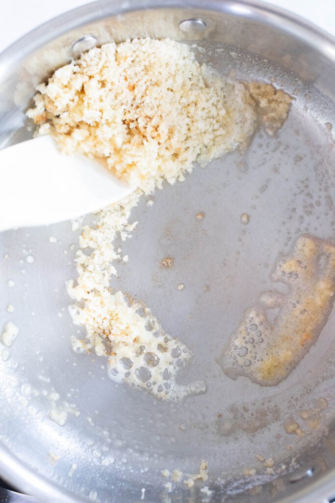 Panko breadcrumbs being toasted in melted butter in a stainless steel pan with a white spatula.