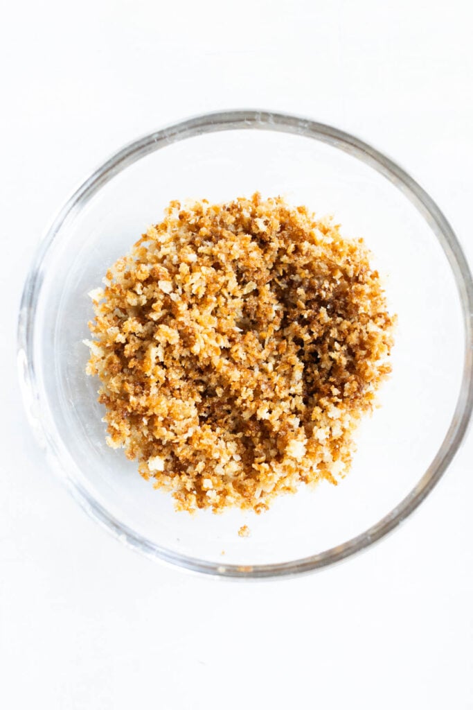 A clear glass bowl containing a mixture of golden brown toasted breadcrumbs on a white surface.