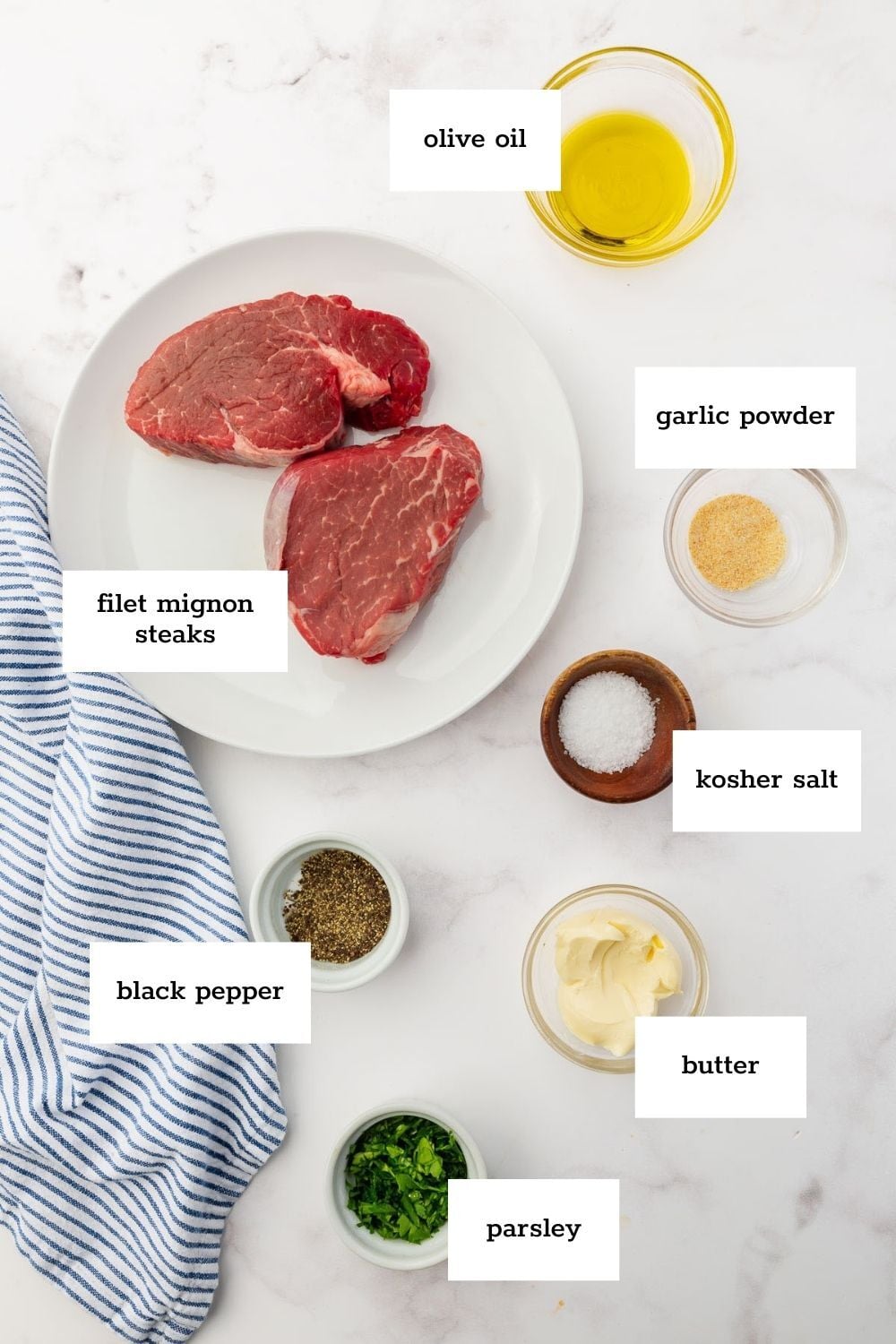 Two raw steaks on a plate, surrounded by bowls of seasoning, butter, oil, salt, and herbs on a white surface with a blue striped towel to the side.
