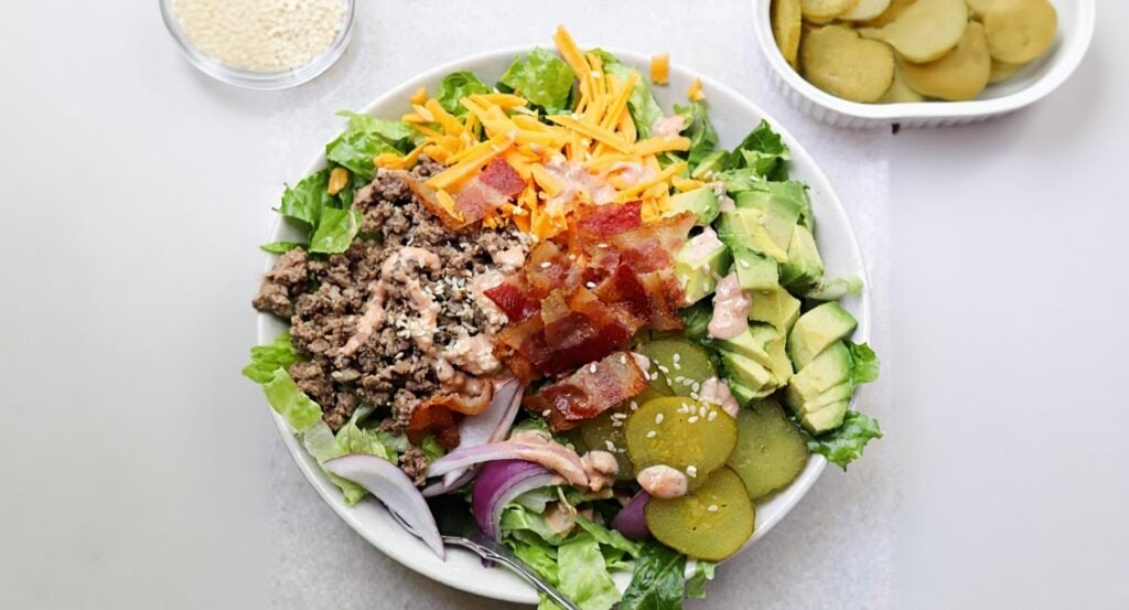 A bowl with ground beef, chopped lettuce, shredded cheddar, bacon, avocado, red onion, pickles, sesame seeds, and dressing. Small bowls of sesame seeds and pickles are beside it.