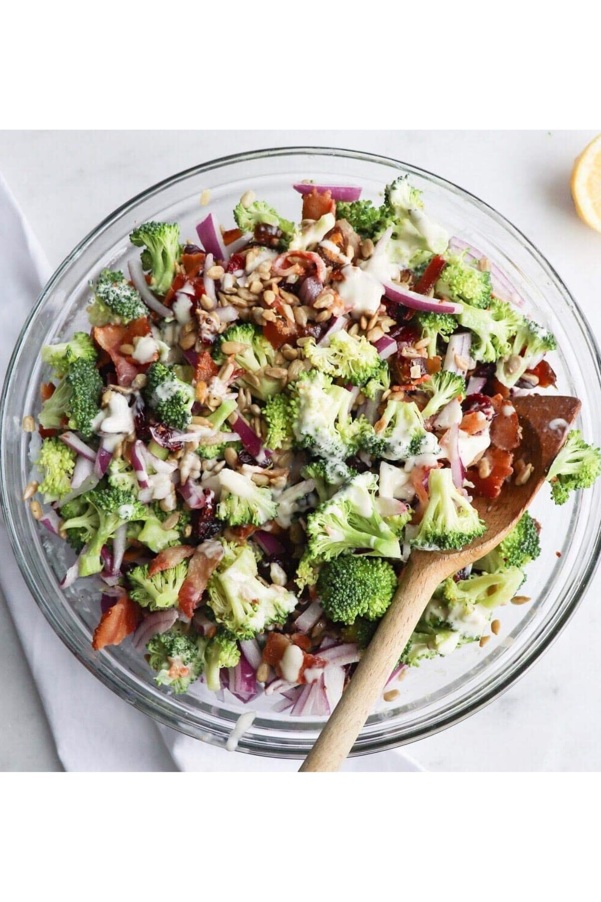 A glass bowl filled with a broccoli salad containing chopped broccoli, red onions, bacon, sunflower seeds, and a creamy dressing, with a wooden spoon resting inside.