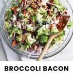 A clear glass bowl filled with chopped broccoli, bacon, red onion, sunflower seeds, and creamy dressing, with a wooden spoon, on a white surface. Text below reads "Broccoli Bacon Salad.