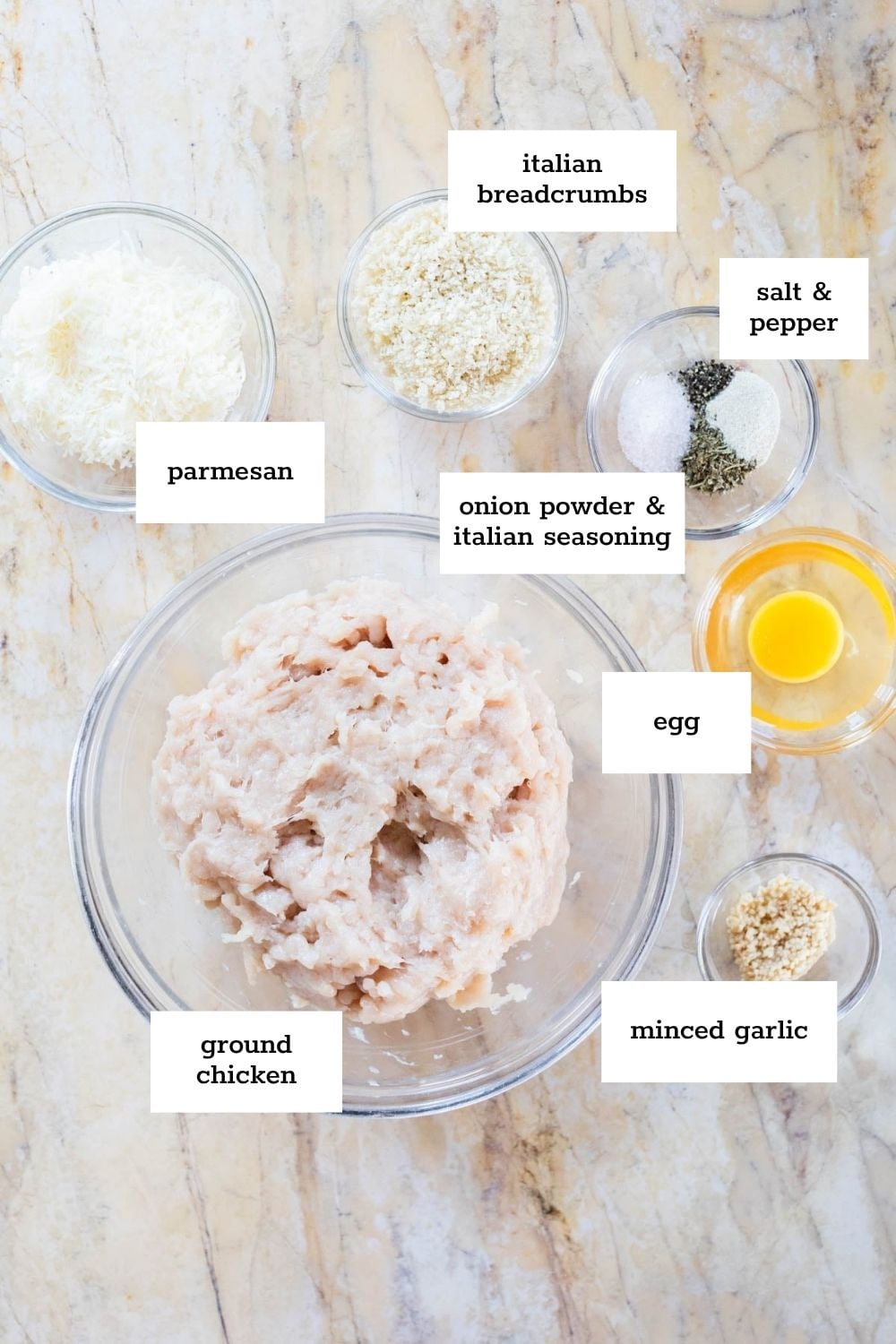 Overhead view of bowls containing ground chicken, minced garlic, egg, onion powder, Italian seasoning, salt, pepper, parmesan, and Italian breadcrumbs on a marble surface.