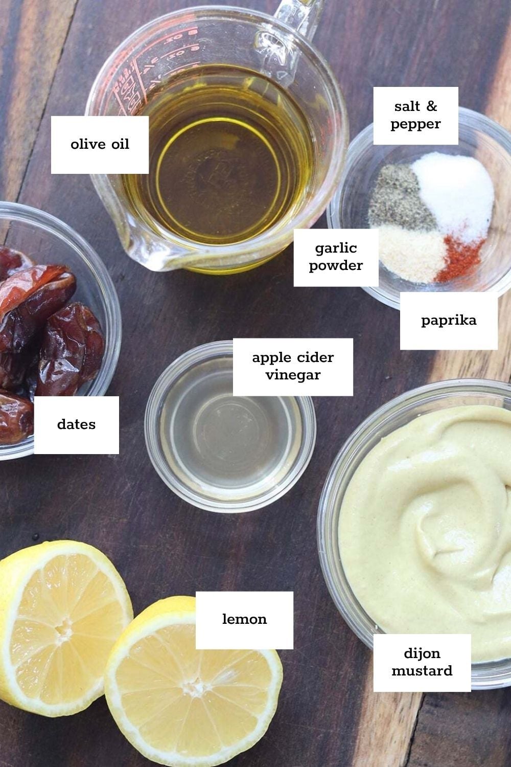 Various labeled ingredients for a dressing or marinade, including olive oil, dates, apple cider vinegar, lemon, dijon mustard, garlic powder, paprika, salt, and pepper on a wooden surface.