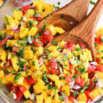 A glass bowl of mango pico de gallo with chopped mango, tomatoes, red onion, cilantro, and jalapeño, mixed with wooden serving utensils—a vibrant salsa fresca recipe perfect for any occasion.